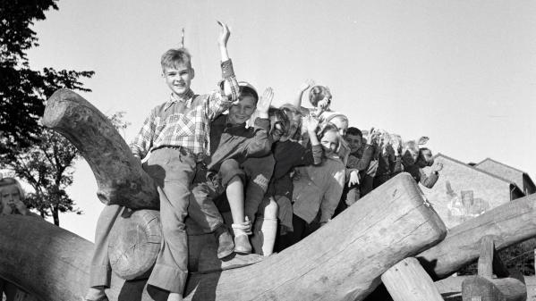 En gammal svartvit bild där en grupp barn sitter på en lekkonstruktion i trä och vinkar.